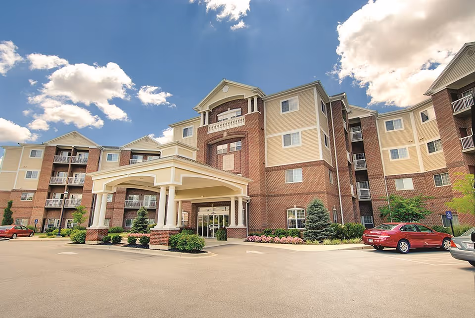Exterior view of a multi-story senior living facility with a covered entrance, brick and beige siding, surrounded by a parking lot with cars and landscaping including small trees and shrubs under a partly cloudy blue sky.