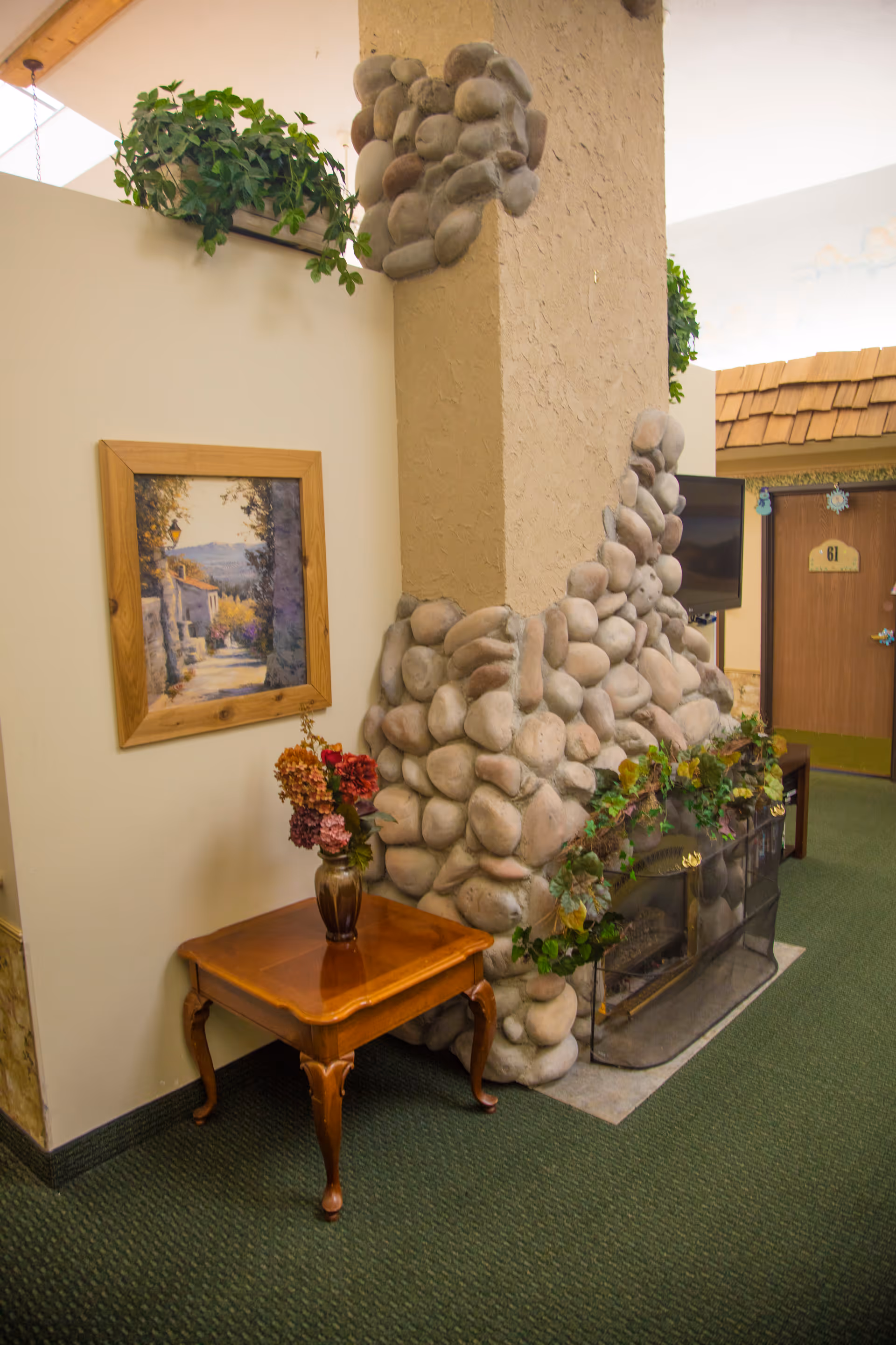 A stone-faced fireplace in an indoor corridor with a wooden side table holding a vase of flowers, a framed painting on the wall, and a room door in the background.