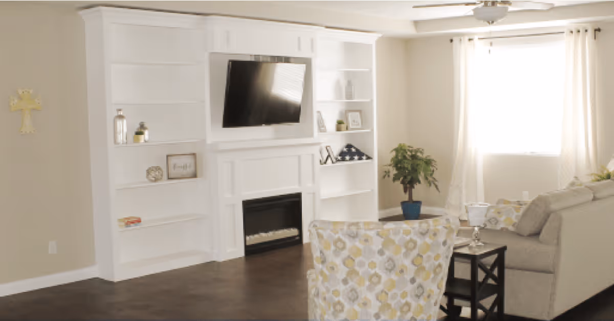 A bright living room with a white built-in shelving unit and fireplace, a mounted flat-screen TV above the fireplace, a patterned armchair, a beige sofa with decorative pillows, a small side table with a lamp, a potted plant, and a window with white curtains letting in natural light.