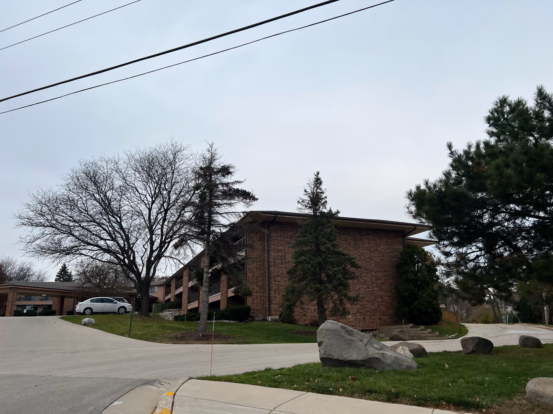Two-story brick building with trees, large rocks, a curved driveway and a parked car under an overcast sky.