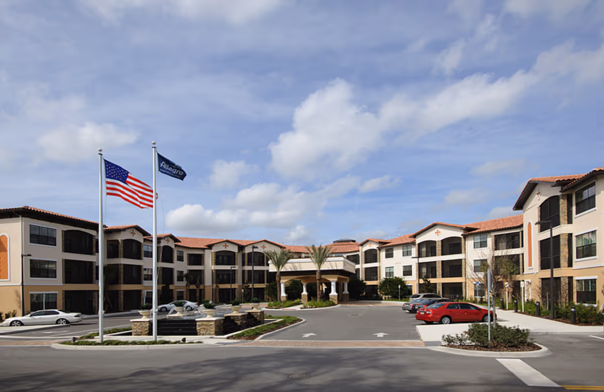 Exterior front view of a three-story senior living facility with American and corporate flags, a circular driveway, and parked cars.