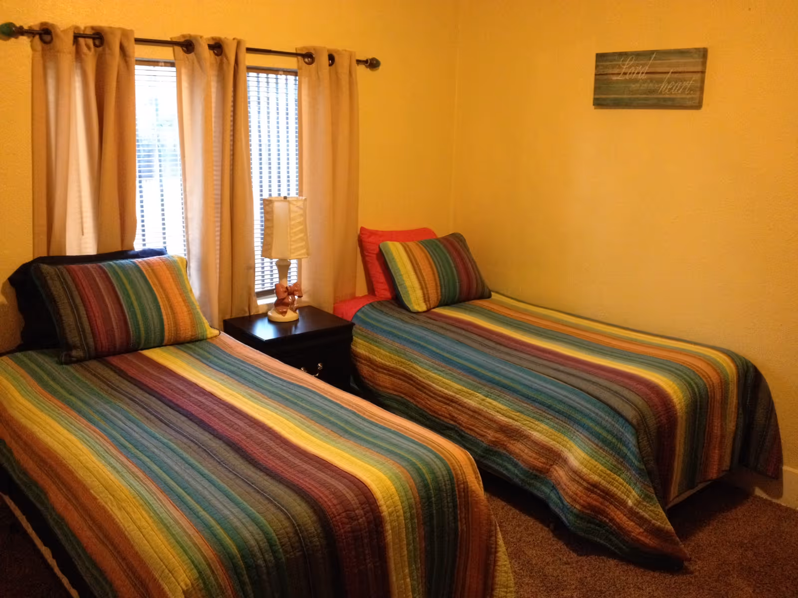 Small bedroom with two twin beds covered in rainbow-striped bedspreads, a nightstand with a lamp between them and a curtained window.