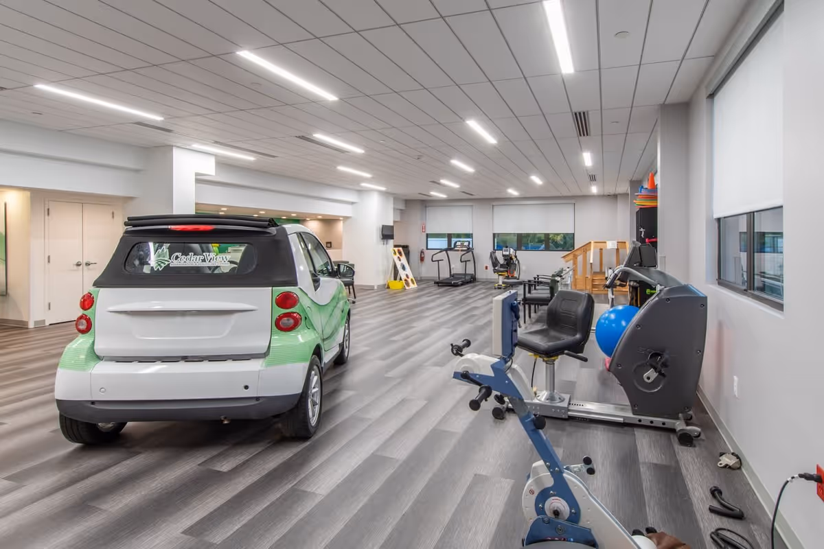 Indoor rehabilitation room with exercise equipment including a stationary bike, treadmill, and other fitness machines. A small green and white car with a Cedar View logo on the rear window is parked inside. The room has large windows with blinds, gray wood-patterned flooring, and bright overhead lighting.