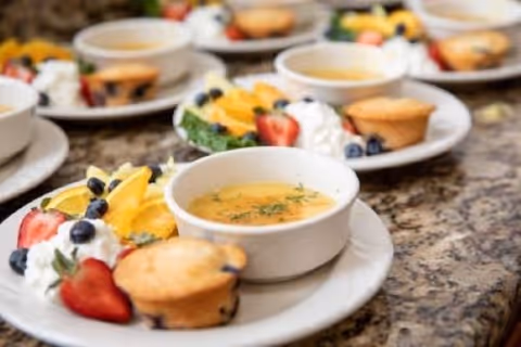 Several white plates arranged on a granite countertop, each plate containing a bowl of soup garnished with herbs, a small muffin, and a side of fresh fruit including strawberries, blueberries, and orange slices with whipped cream.