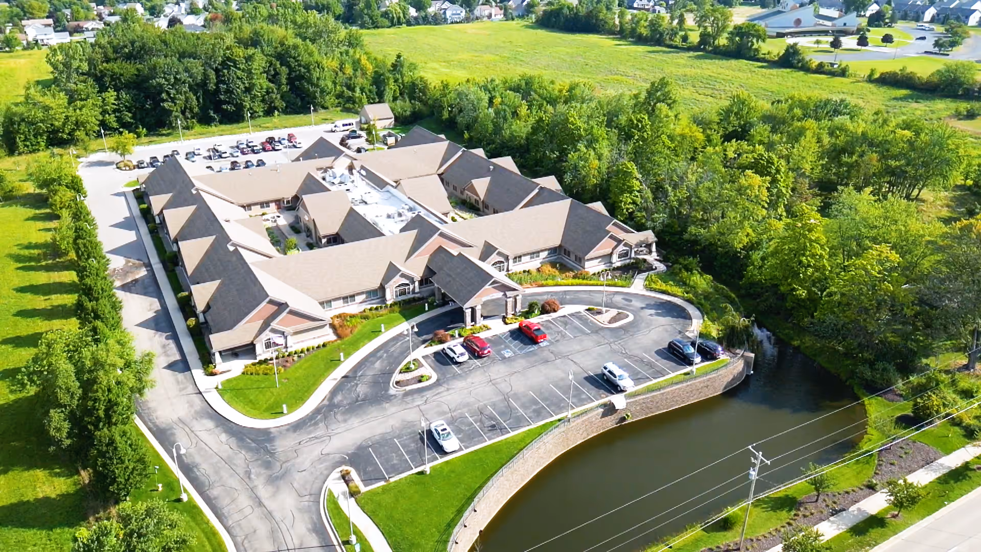 Aerial view of Medilodge of Holland facility surrounded by greenery and trees. The building has a beige roof and is shaped in a U-formation with a parking lot in front containing several cars. There is a small body of water adjacent to the parking lot and a road running alongside the property.