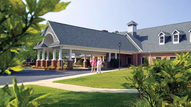 Exterior view of a senior living facility building with a covered entrance and a paved driveway. Two elderly women are walking on the sidewalk near the entrance. The building has a brick facade, white columns, and a gray shingled roof with dormer windows. Green shrubs and trees surround the area.