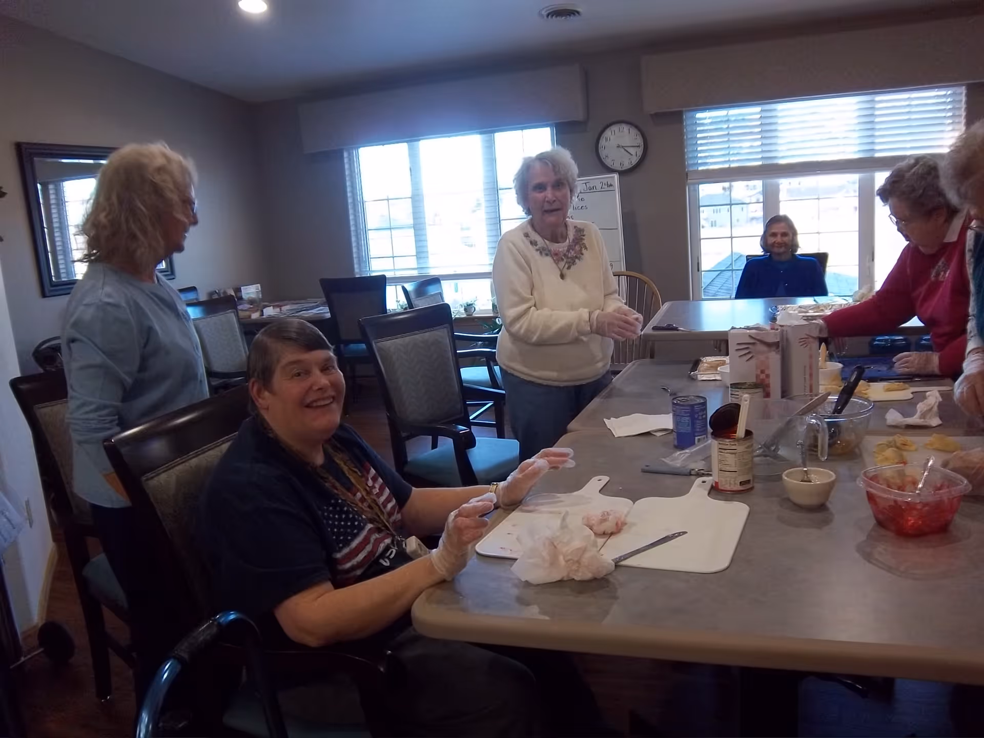 A group of elderly residents prepare food together around a large table in a senior living common room.