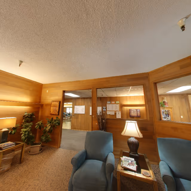 A cozy interior room with wood-paneled walls and a textured ceiling. The room features two blue upholstered armchairs separated by a small wooden side table with a lamp and magazines. There are potted plants on the left side and a windowed wall showing an adjacent room with bulletin boards and more wood paneling.