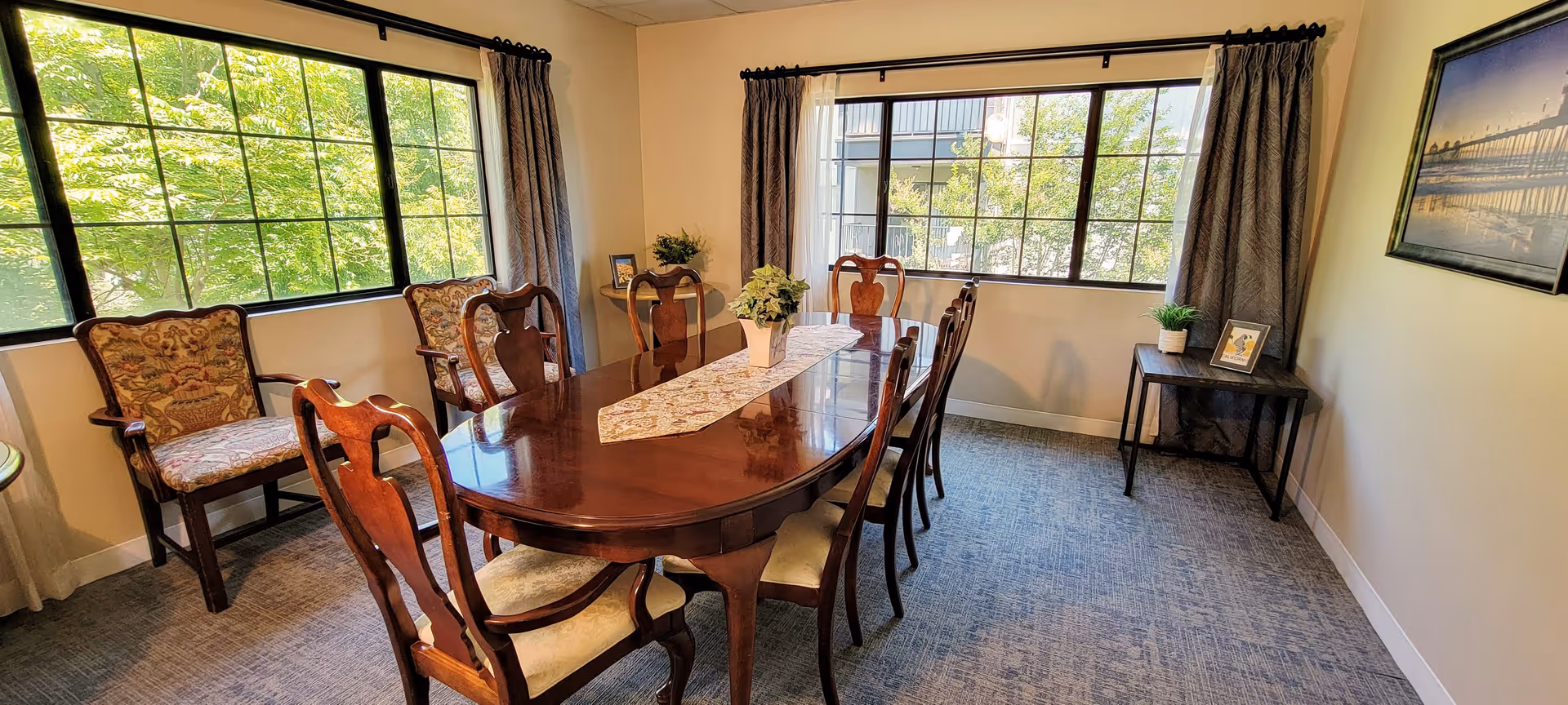 A well-lit dining room with a polished wooden dining table surrounded by eight chairs. The room has large windows with dark curtains allowing natural light to fill the space. There are two armchairs with patterned upholstery near one window, a small side table with a framed photo and a potted plant, and a framed picture hanging on the wall.