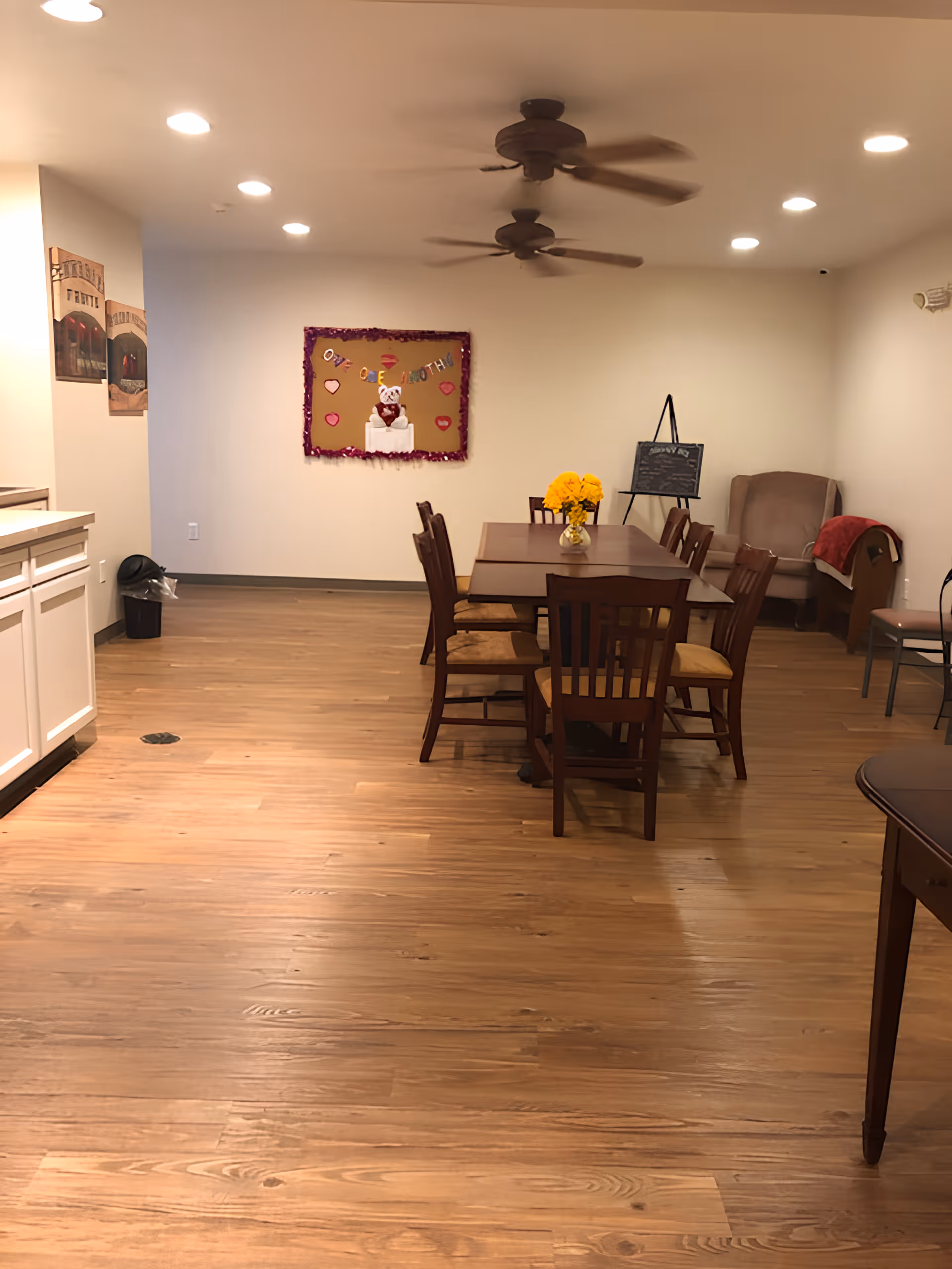 A senior living facility common area with a wooden dining table and chairs in the center. There are two ceiling fans above, a bulletin board decorated with hearts and a teddy bear on the back wall, and a comfortable armchair with a red blanket in the corner. The floor is wooden, and the walls are painted white with some framed pictures on the left side.
