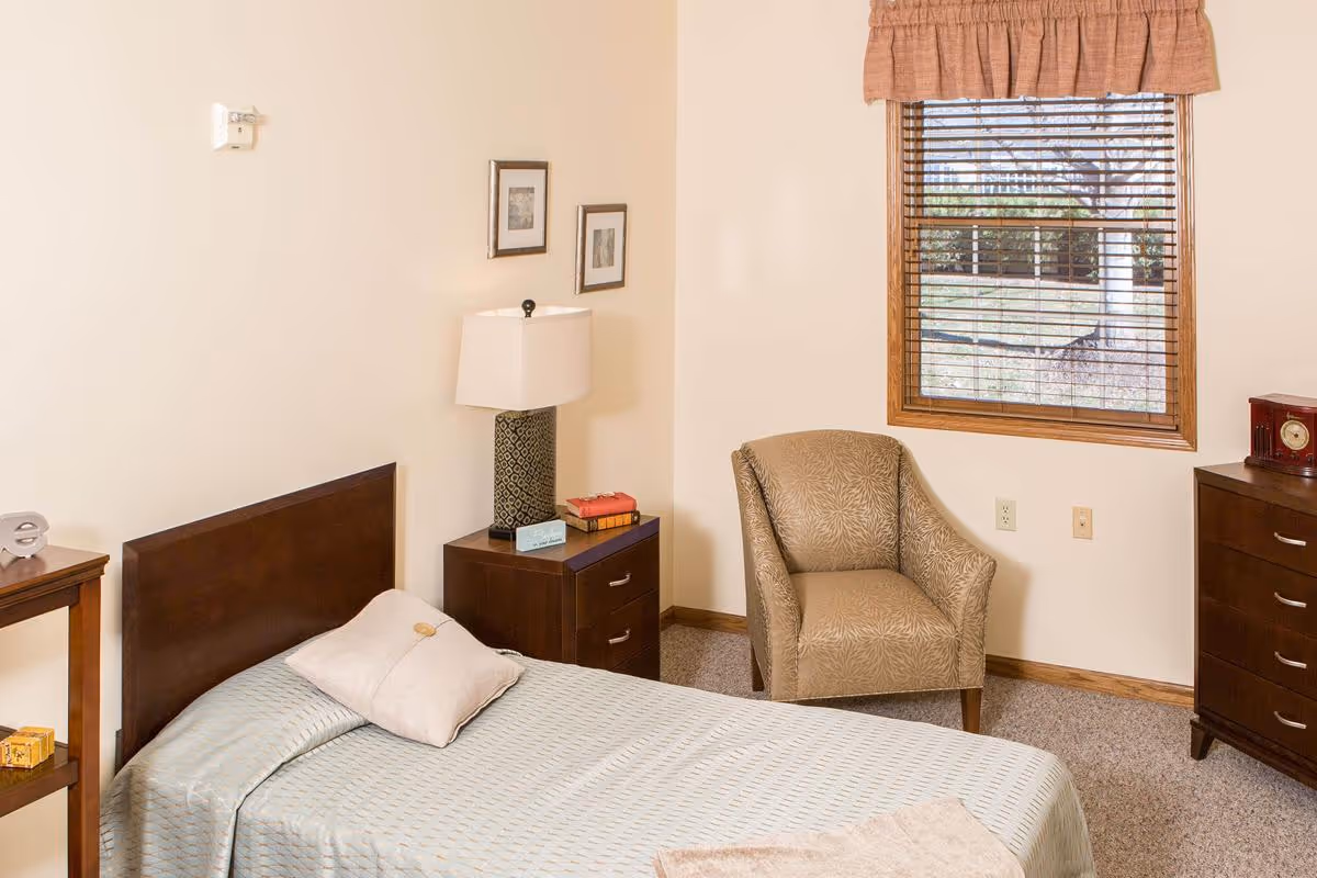 A cozy bedroom with a single bed covered in a light blue bedspread and a beige pillow. Next to the bed is a dark wooden nightstand with a lamp and some books. There is a beige patterned armchair near a window with wooden blinds and a valance. The room has light-colored walls and carpeted floor, with a small dresser and a clock on it.