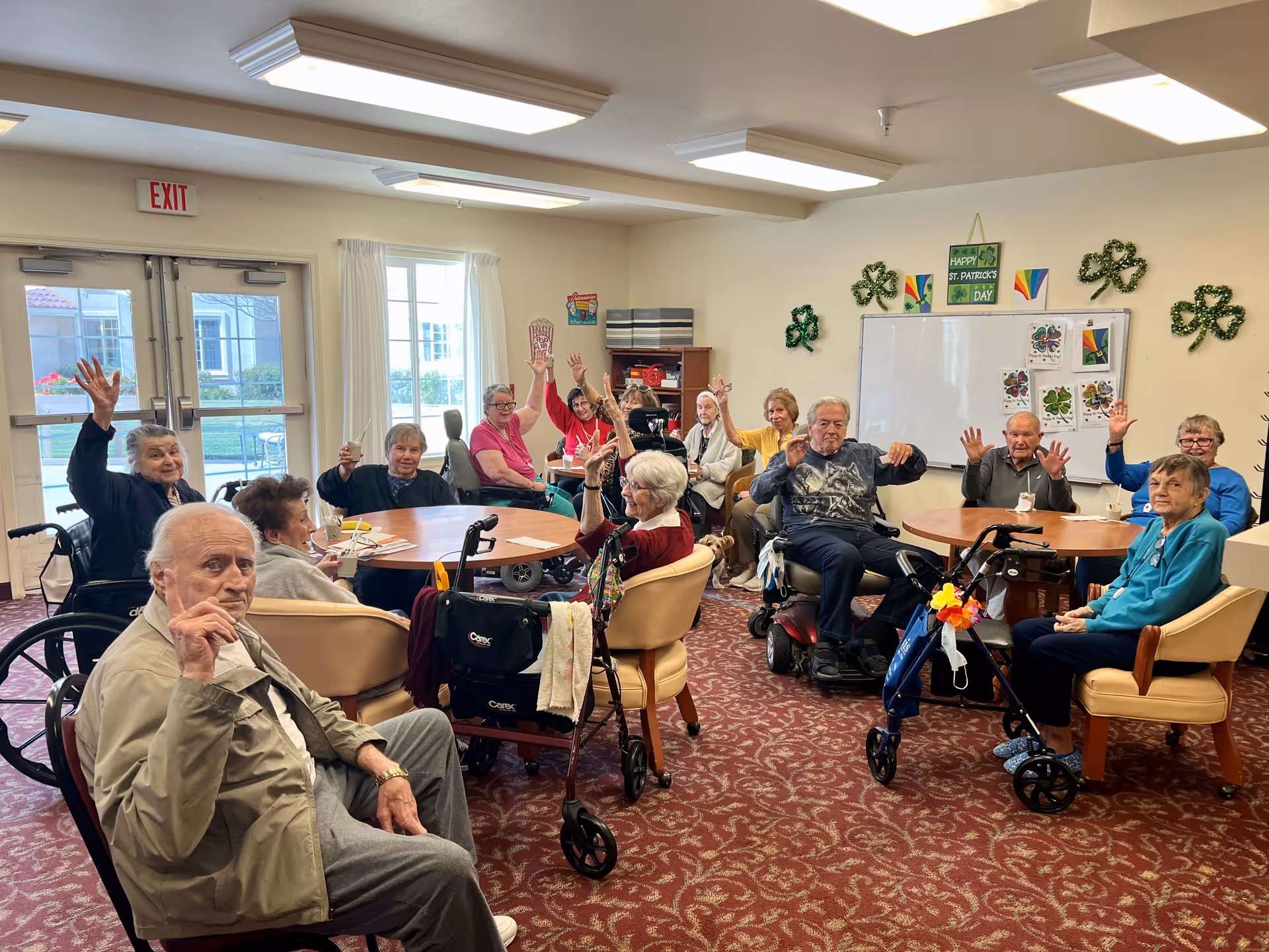 A group of elderly people sitting around tables in a community room, some in wheelchairs and others in chairs, raising their hands and smiling. The room is decorated with shamrocks and St. Patrick's Day decorations on the walls. There is a large window and double doors letting in natural light.