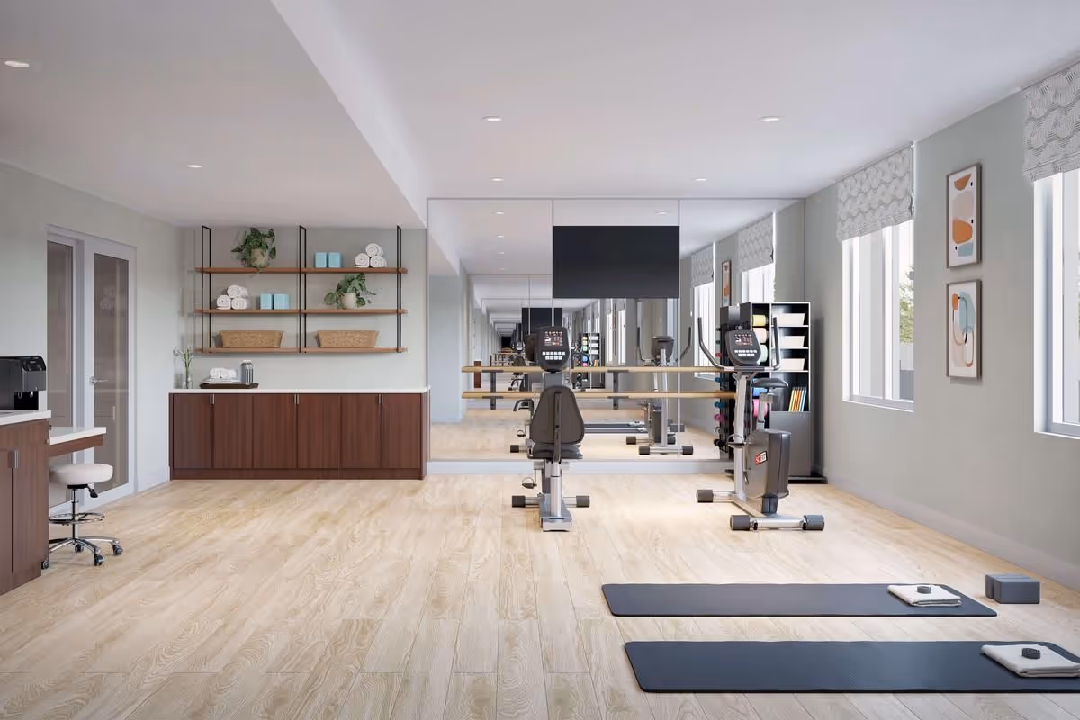 A bright and spacious fitness room with light wood flooring, two exercise mats with towels and weights, two stationary exercise bikes facing a large wall mirror, shelves with towels and plants, a TV mounted on the mirror, and windows with patterned blinds letting in natural light.