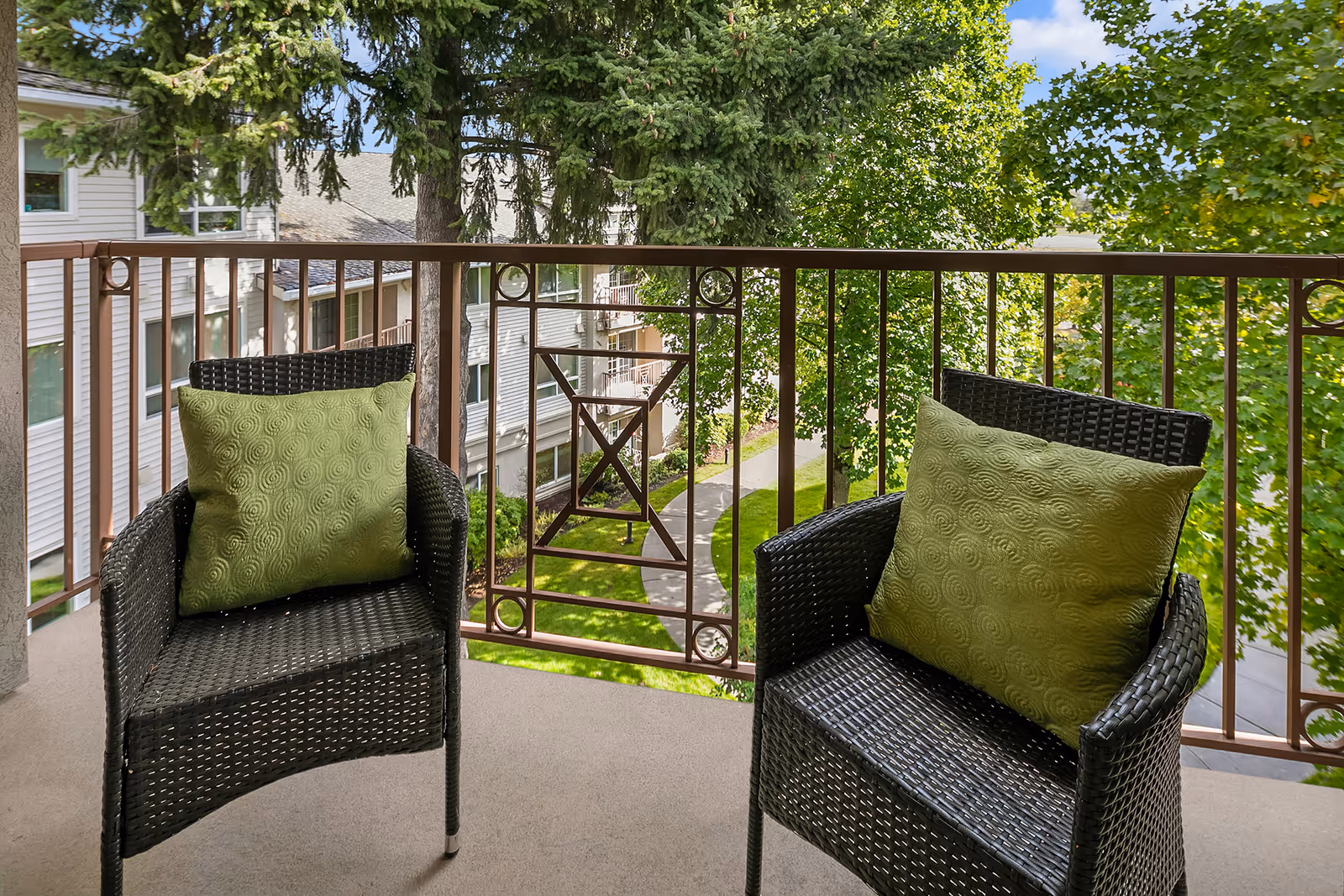 Two black wicker chairs with green cushions on a balcony overlooking a pathway, trees, and parts of a residential building.