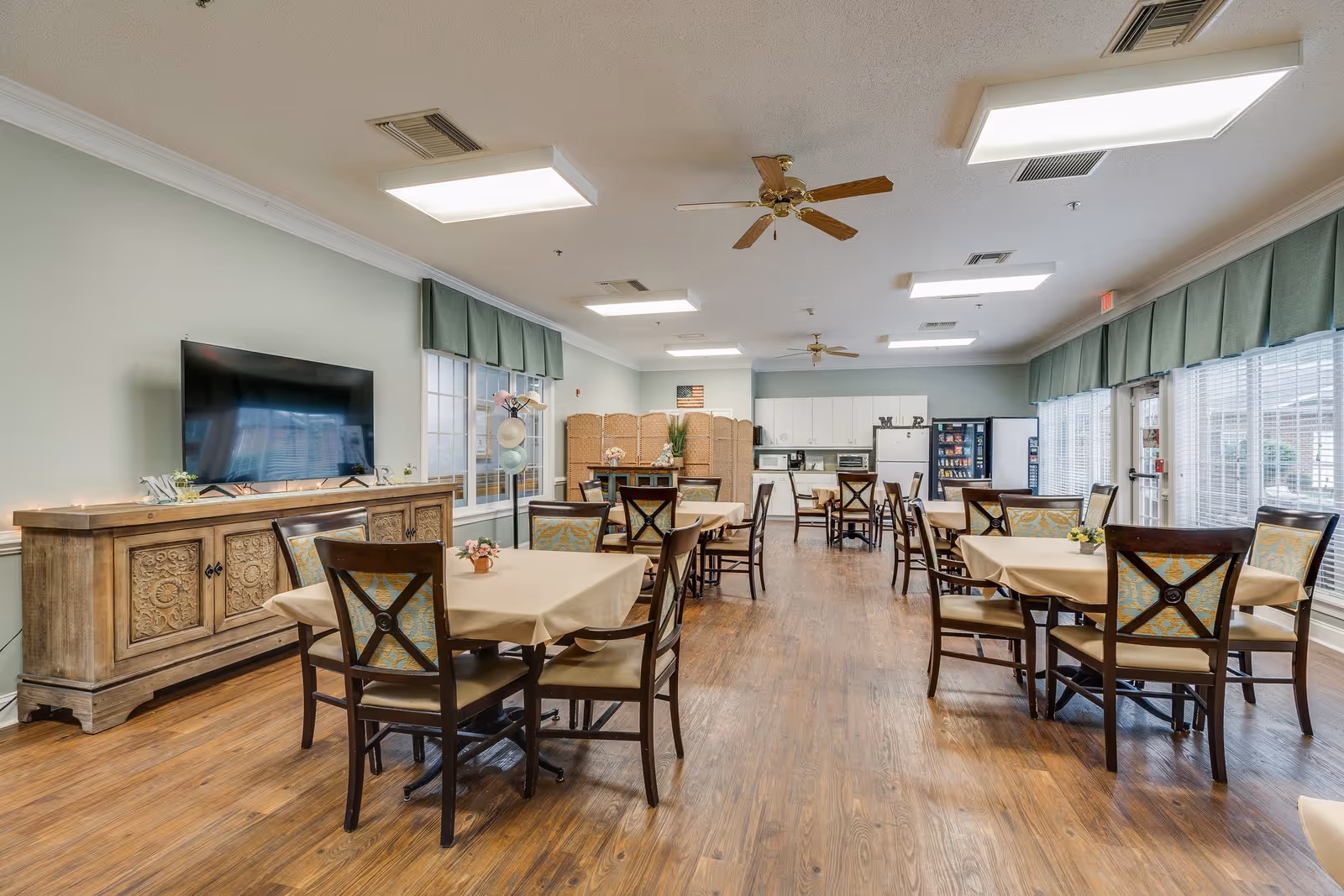 Bright communal dining room with multiple tables and chairs, a TV, and a kitchenette in the background.