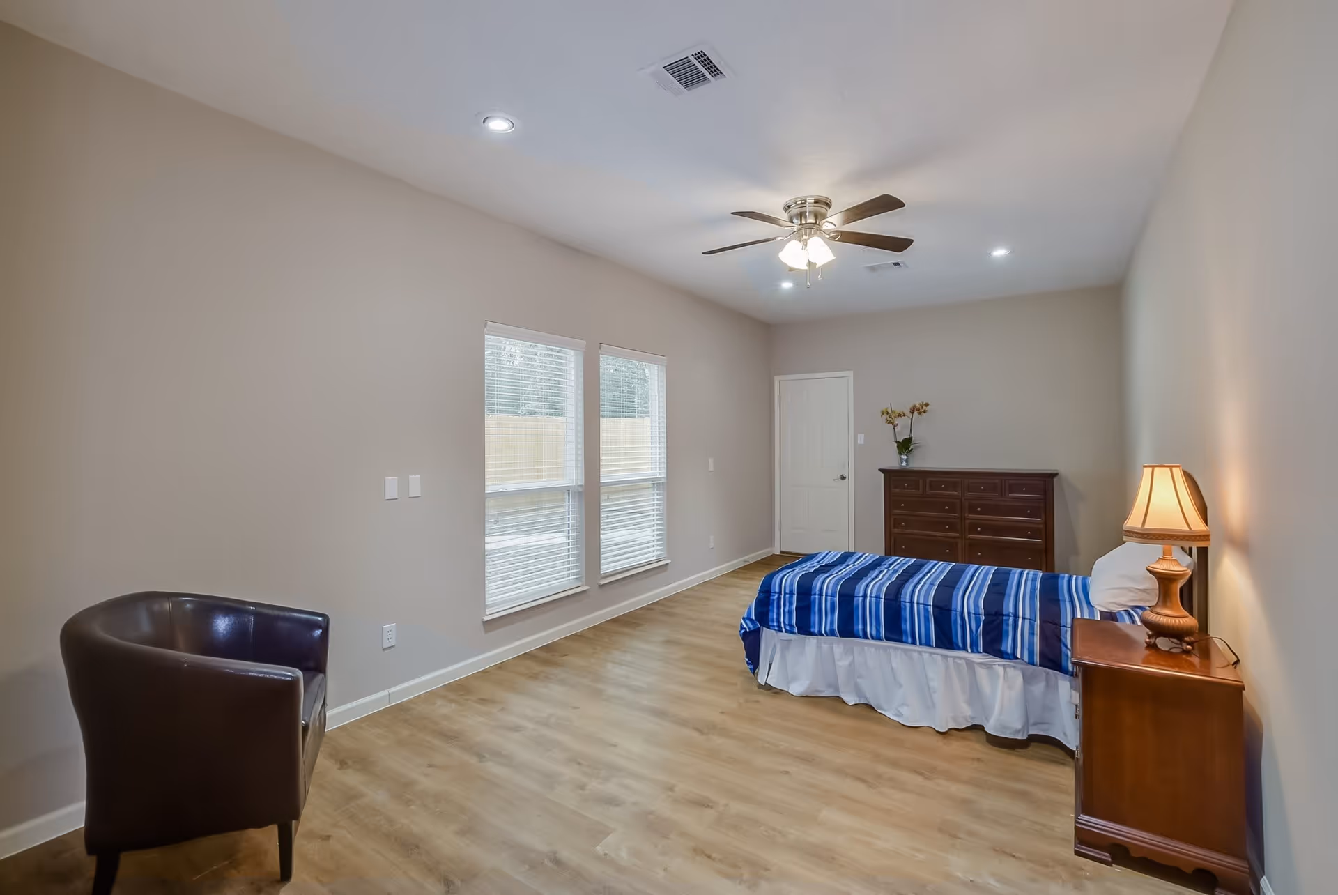 A simple bedroom with light beige walls and wood flooring. The room contains a single bed with a blue striped blanket, a wooden nightstand with a lamp, a wooden dresser with a vase of flowers, and a dark brown armchair. Two windows with white blinds allow natural light into the room. A ceiling fan with lights is mounted on the ceiling.