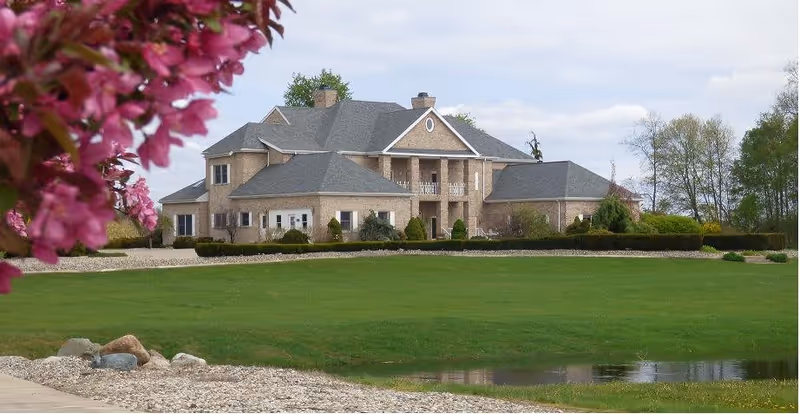 Large two-story brick building with multiple roof sections and a front porch, surrounded by green lawn and trees, with pink flowers in the foreground on the left side.