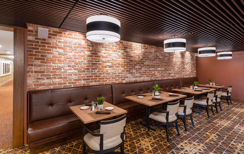 Cozy dining room with booth seating along an exposed brick wall, wooden tables and chairs, and pendant lights overhead.