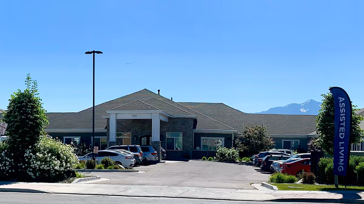 Front exterior view of Sunridge Assisted Living and Memory Care facility with a parking lot in front, several parked cars, landscaped bushes and trees, a tall street lamp, and a blue flag sign that reads 'ASSISTED LIVING'. Mountains are visible in the background under a clear blue sky.