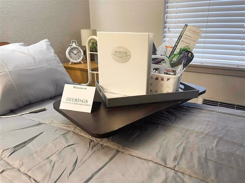 A neatly made bed with gray sheets and pillows in a room. On the bed is a dark wooden overbed table holding a tray with a welcome card, a folder labeled Deerings Nursing & Rehabilitation, and a basket containing various items. A small white alarm clock and a potted plant are on a wooden nightstand next to the bed. A window with closed blinds is in the background.