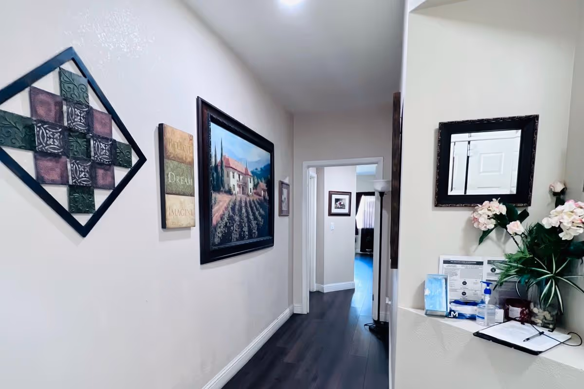 A hallway in an elderly care facility with framed artwork on the left wall and a small counter on the right holding a vase with flowers, hand sanitizer, and some documents. The hallway has dark wood flooring and leads to a room with a window covered by curtains.