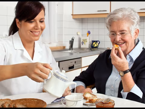 A caregiver pouring tea into a cup while an elderly woman smiles and eats a piece of bread with jam in a kitchen setting.