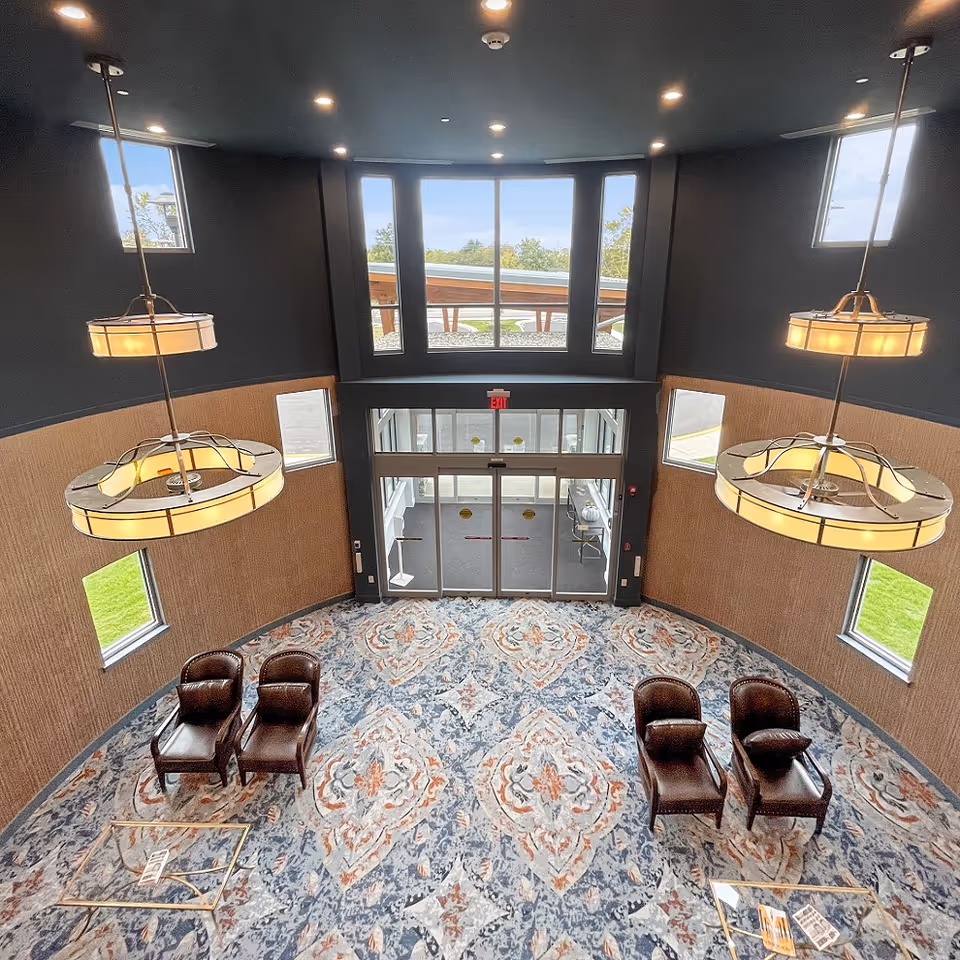 View from above of a spacious lobby area with large windows letting in natural light. The room features a patterned carpet with blue, orange, and white colors, four brown leather chairs arranged in pairs, two modern hanging light fixtures, and a glass entrance door with an exit sign above it.