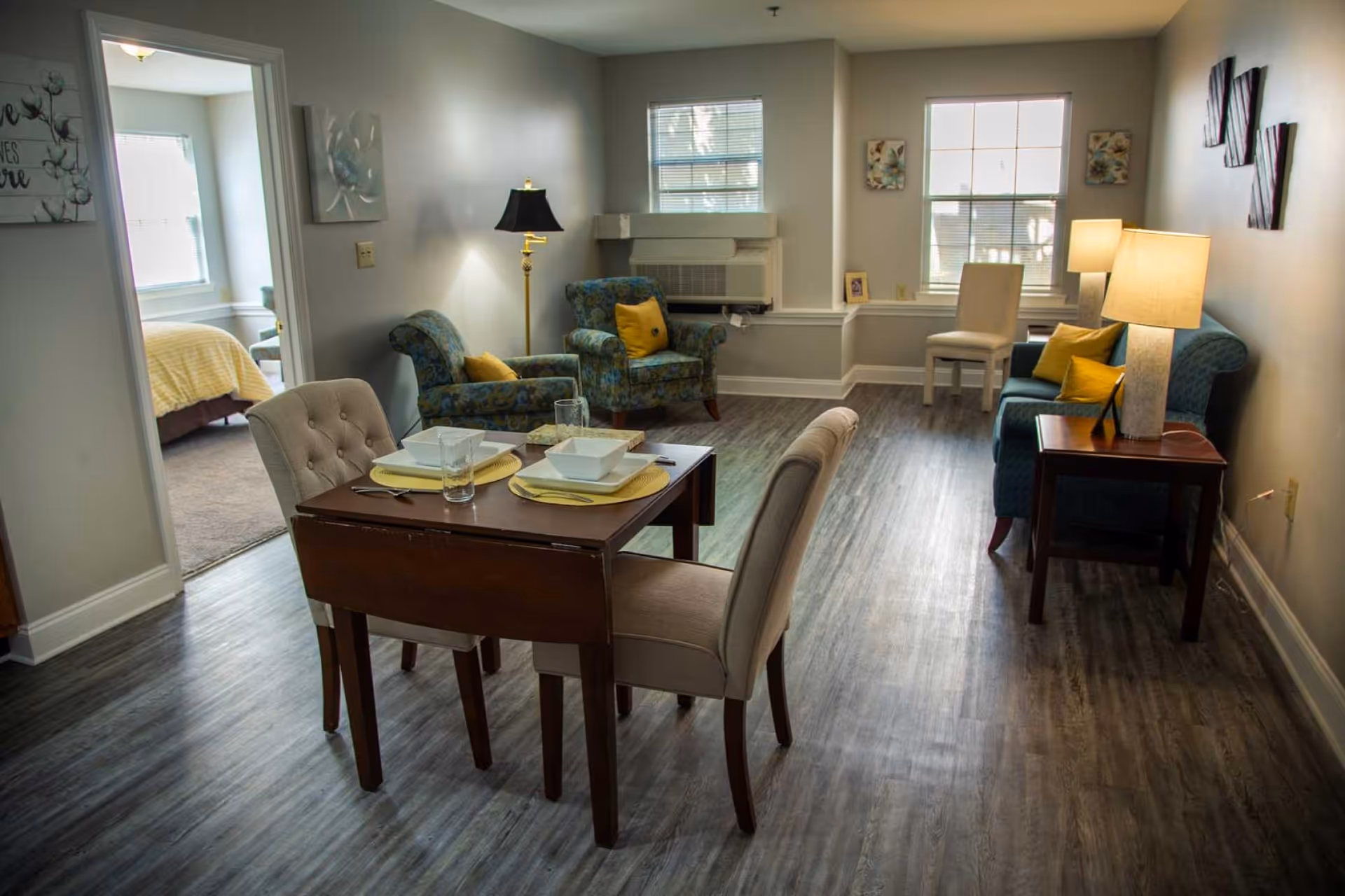A cozy living and dining area in a senior living facility with a small wooden dining table set for two, two upholstered armchairs with yellow cushions, a blue loveseat with yellow cushions, a side table with a lamp, and two windows letting in natural light. A bedroom with a bed covered in a yellow blanket is visible through an open doorway.
