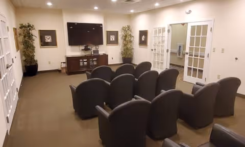 A small theater-style room with two rows of black cushioned chairs facing a wall-mounted flat screen TV. The room has beige walls, carpeted floor, two tall potted plants on either side of the TV, framed artwork on the walls, and double glass doors on the right side.