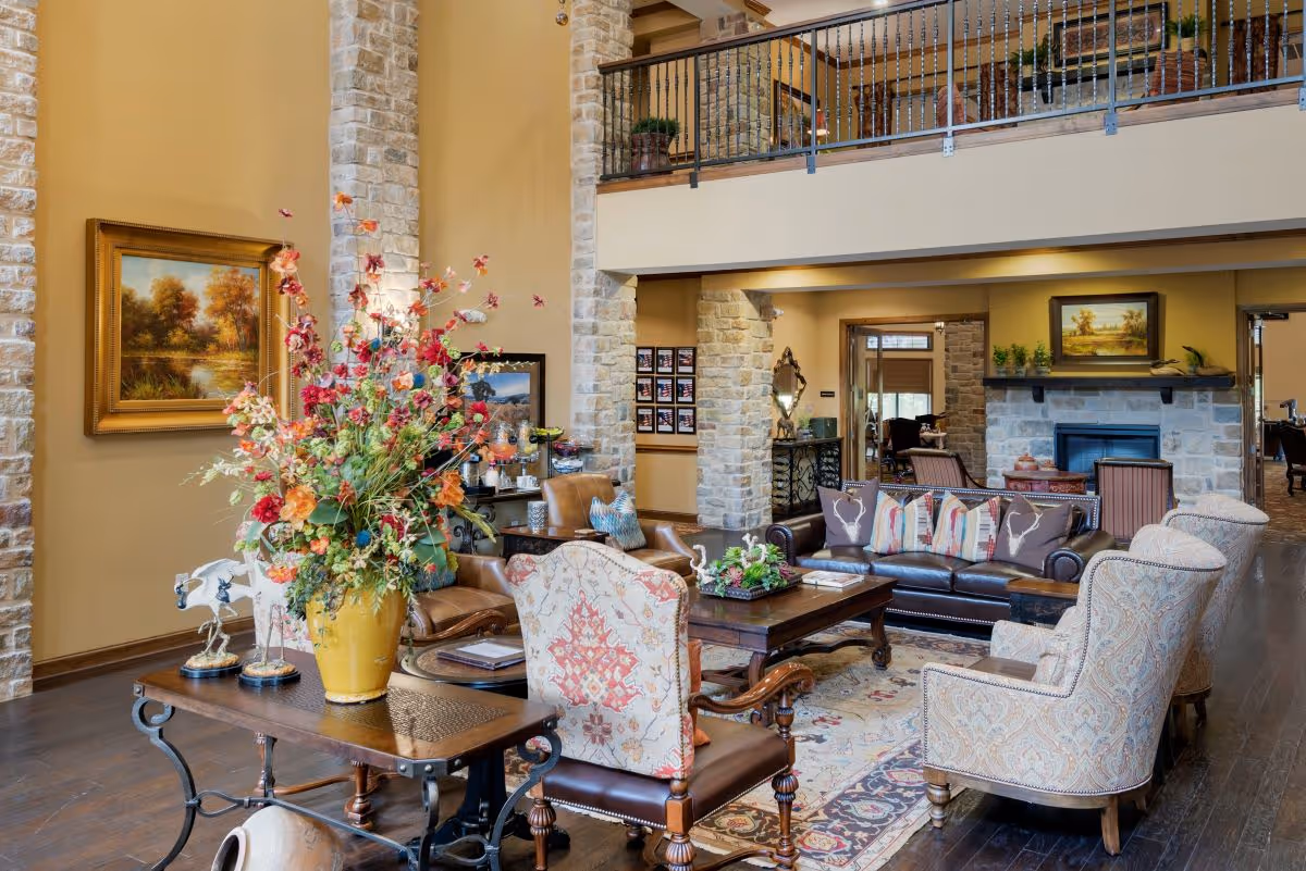 Spacious, elegantly furnished common living room with high ceilings, stone columns, multiple armchairs and sofas arranged around a coffee table and fireplace.