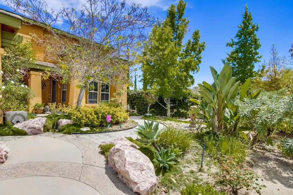 A sunny outdoor garden area at Silver Oaks, South featuring a curved concrete pathway surrounded by large rocks, various green plants, bushes, and trees. A yellow building with windows and a door is visible in the background under a clear blue sky.