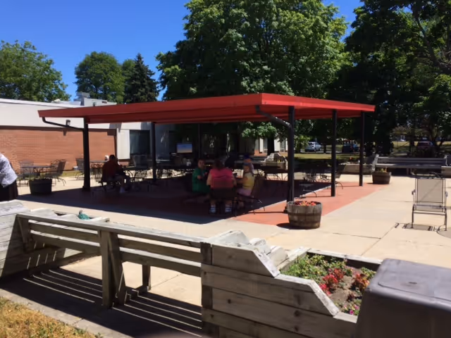 Outdoor seating area at a senior living facility with a red canopy providing shade over several tables and chairs. People are seated under the canopy, and there are trees and a brick building in the background. Wooden benches and planters with flowers are visible in the foreground.