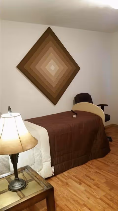 A simple bedroom with a single bed covered in a brown quilt and a white blanket at the foot. There is a wooden nightstand with a lamp on it next to the bed. A dark office chair is positioned near the head of the bed. On the wall above the bed hangs a large square piece of art with concentric diamond shapes in varying shades of brown. The floor is wooden, and the walls are plain white.