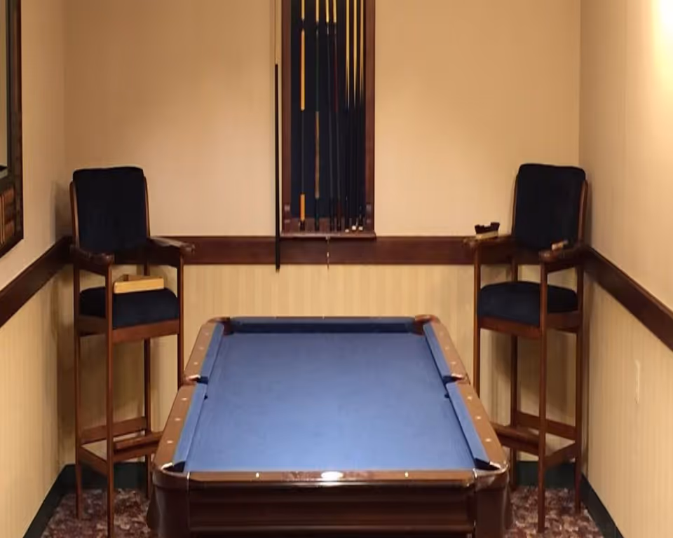 A small room with a blue felt pool table centered between two tall wooden chairs with dark blue cushions. A rack holding pool cues is mounted on the wall behind the table.