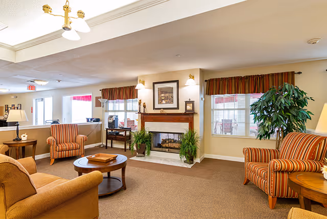 A cozy living room area in a senior living facility featuring a fireplace with framed artwork above it, two striped armchairs, a beige sofa, a round wooden coffee table, side tables with lamps, and large windows with valances. There are also green plants placed near the fireplace and window.
