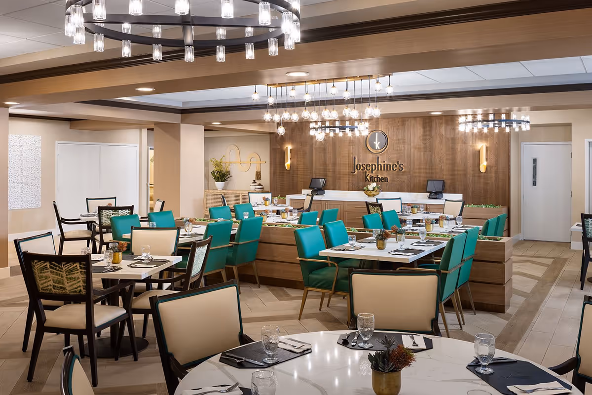 A modern dining room with multiple tables set with glasses, napkins, and cutlery. The chairs are a mix of teal and beige upholstery. The room features contemporary chandeliers and a wooden accent wall with the sign 'Josephine's Kitchen'.