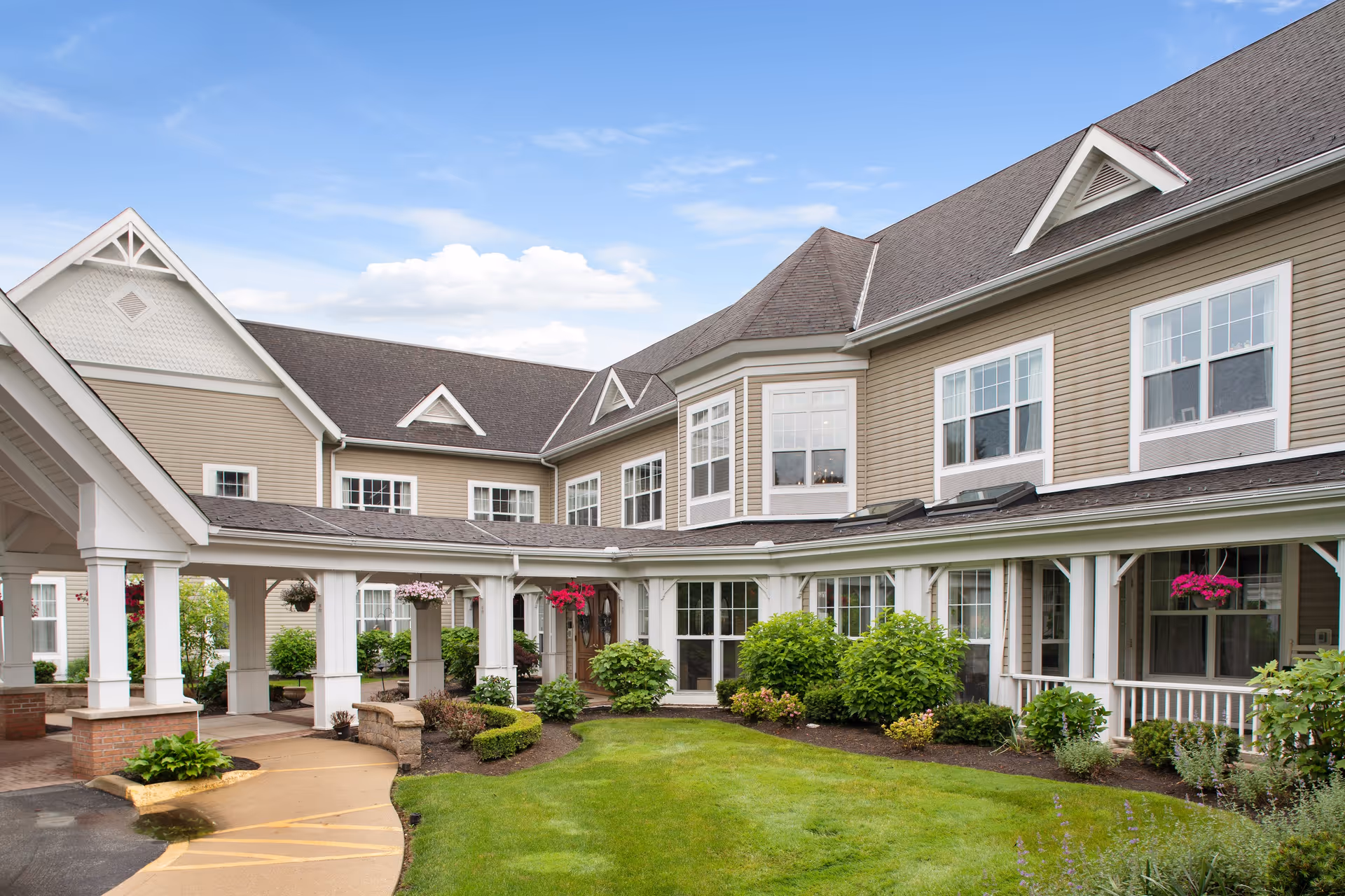Front exterior of a two-story senior living facility with a covered entrance, landscaped lawn, and hanging flower baskets.