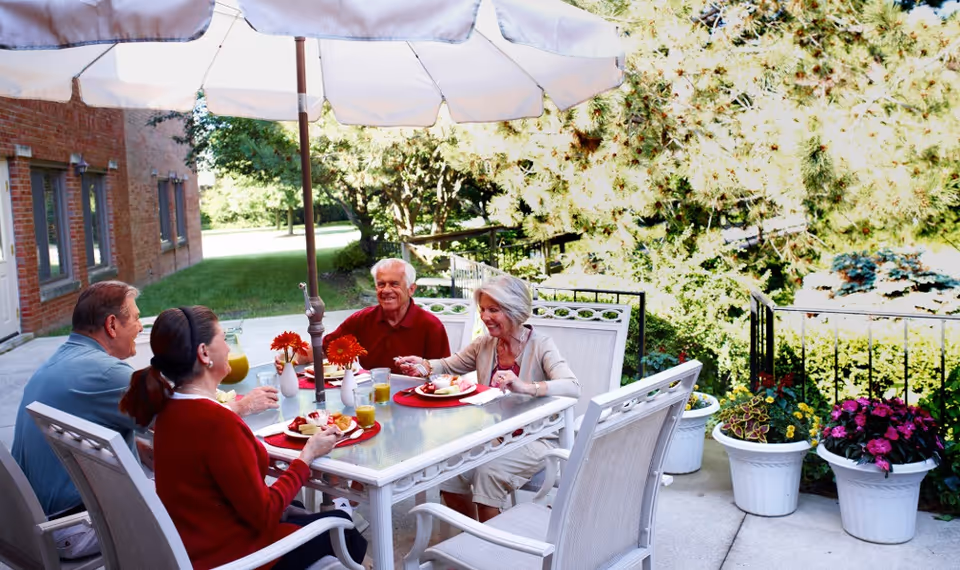 Four elderly people sitting around a white outdoor table with an umbrella, enjoying a meal together on a patio surrounded by greenery and potted flowers.