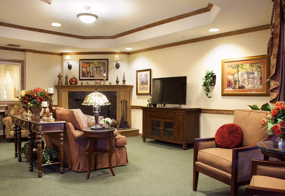 A cozy living room in a senior living facility featuring a patterned sofa and armchair with a red round pillow, a wooden TV stand with a flat-screen TV, a wooden console table with decorative items and flowers, a fireplace with a mantel decorated with small sculptures and framed artwork above it, and warm lighting from ceiling fixtures and wall sconces.