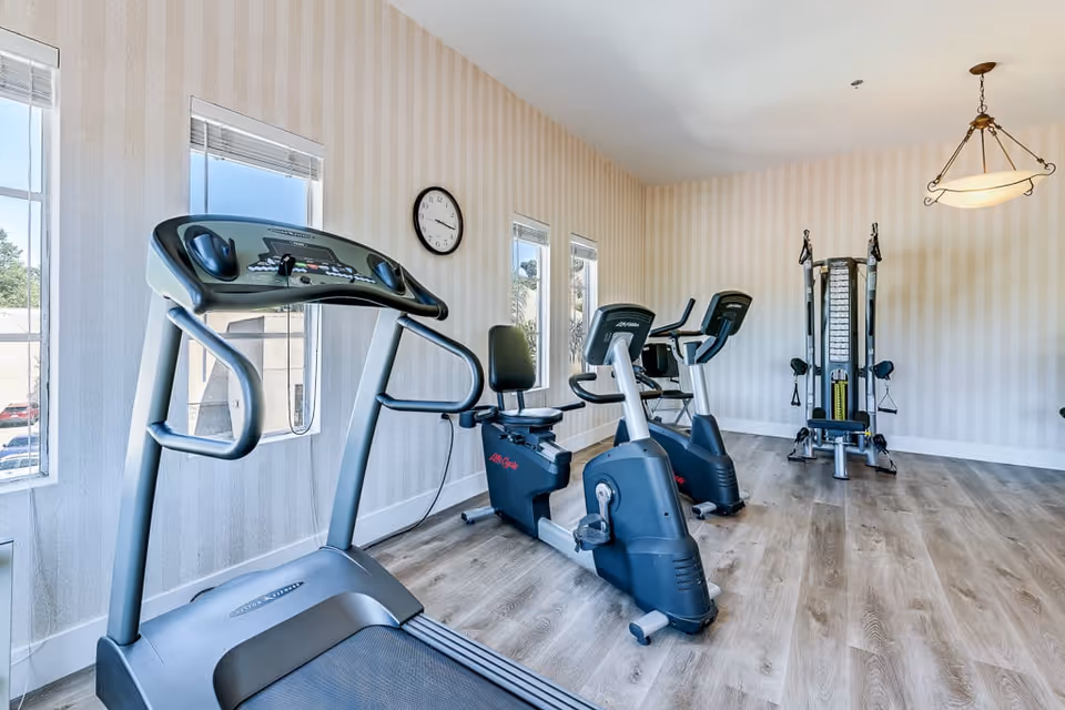A bright exercise room with light wood flooring and beige striped wallpaper. The room contains a treadmill, two stationary bikes, and a multi-function weight machine. Three windows allow natural light to enter, and a round clock is mounted on the wall. A hanging light fixture is visible on the ceiling.