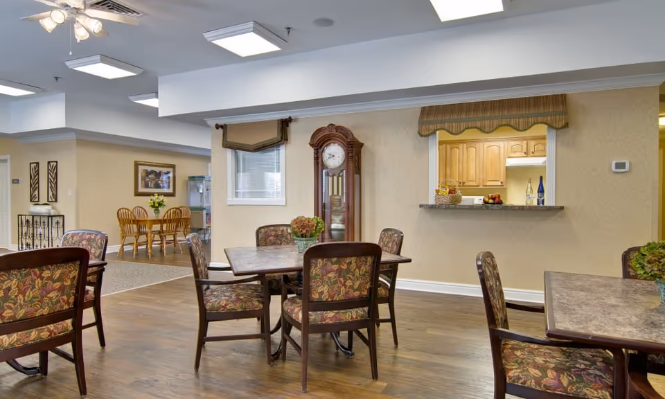 Interior view of a senior living facility common area with multiple tables and chairs featuring floral upholstery. A grandfather clock stands against the wall near a window with a valance. There is a pass-through window to a kitchen area with wooden cabinets and a countertop displaying fruit and bottles. The flooring is wood, and the walls are beige with crown molding. In the background, there is another dining area with a wooden table and chairs and a framed picture on the wall.