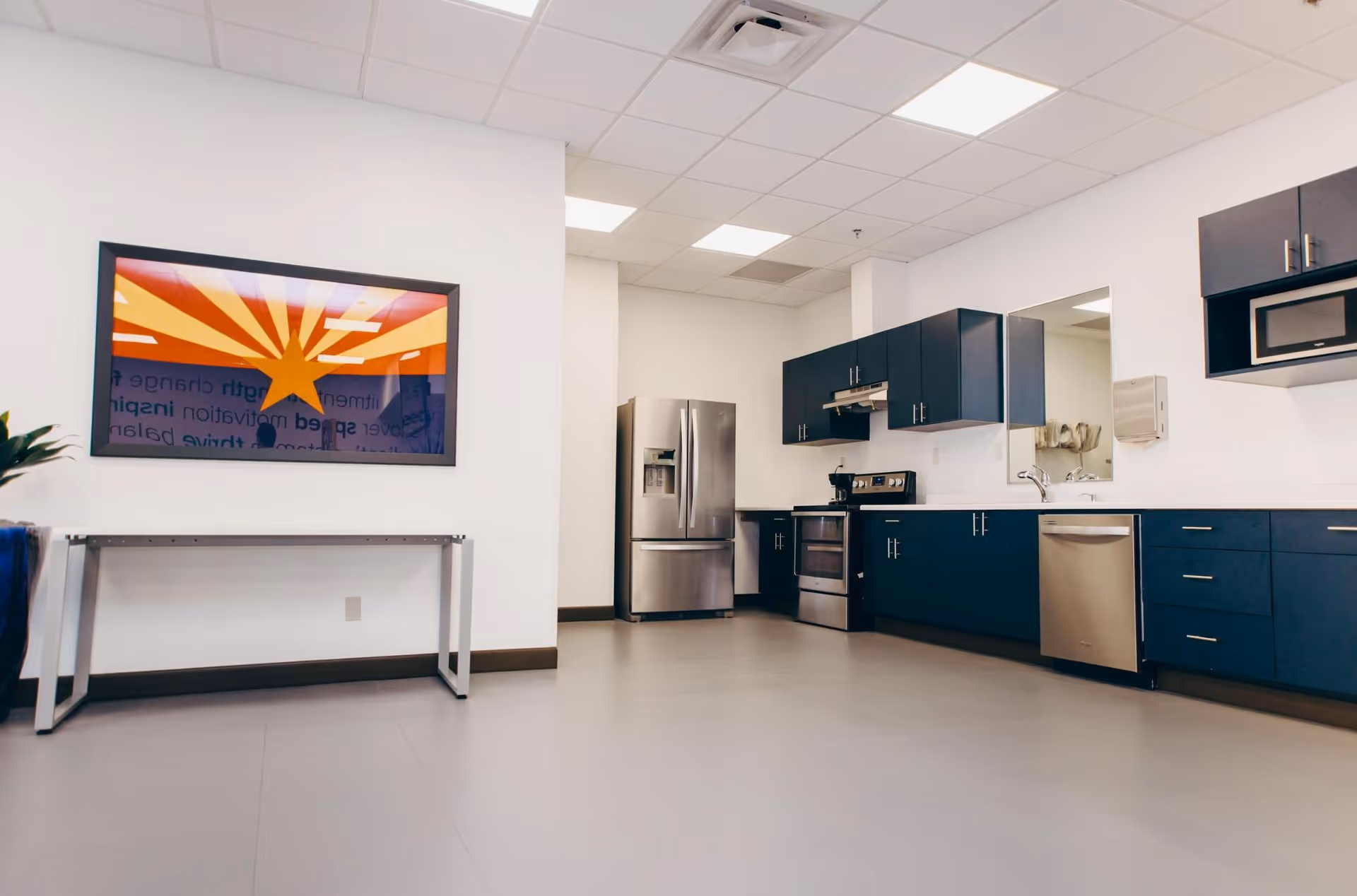 Modern kitchen area with stainless steel refrigerator, stove, dishwasher, and microwave. Blue cabinets with silver handles line the walls. A framed artwork featuring a star and rays in red, yellow, and orange hangs on a white wall above a metal table. The floor is light gray, and the ceiling has recessed lighting panels.