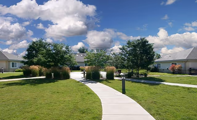 A paved walkway curves through a well-maintained grassy courtyard with trees and ornamental grasses, surrounded by single-story buildings under a partly cloudy blue sky.