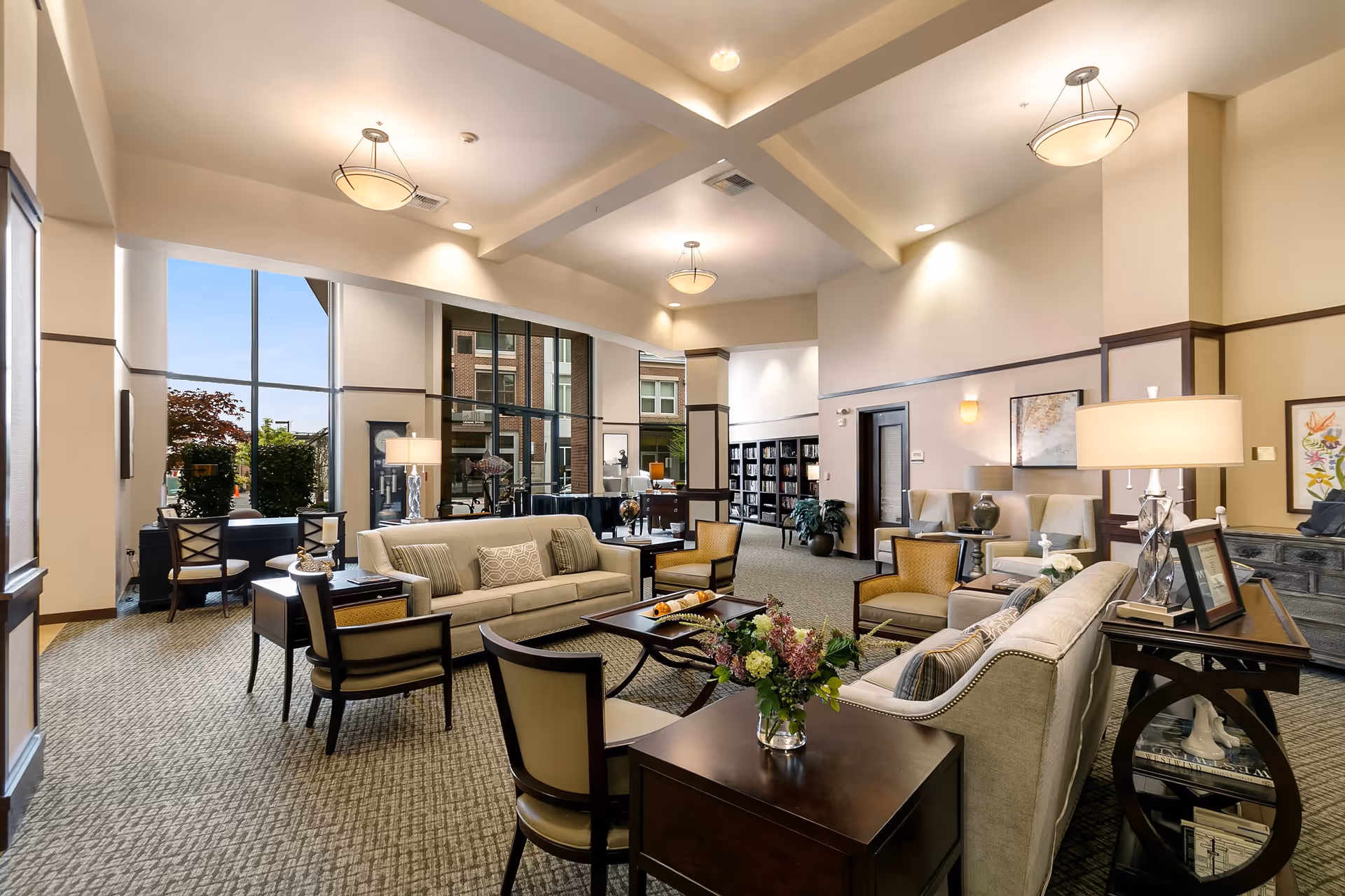 A spacious and well-lit living room area in a senior living facility featuring beige sofas, armchairs, coffee tables with floral arrangements, and large windows allowing natural light. The room has high ceilings with modern light fixtures, carpeted floors, and decorative artwork on the walls. There is a bookshelf and additional seating in the background.