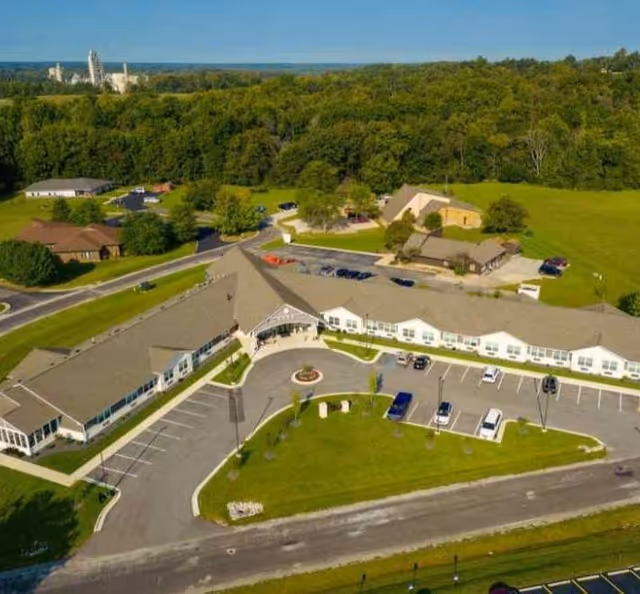 Aerial view of Autumn Trace Kentland Assisted Living facility showing a large, single-story building with a U-shaped layout surrounded by parking lots and green lawns, with trees and other buildings in the background.