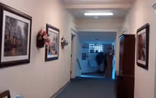 A hallway in a senior living facility with framed paintings and floral wall decorations on the walls. The hallway leads to a room with exercise equipment visible in the background.