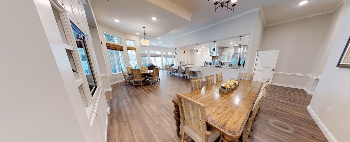 A spacious, well-lit dining area with wooden floors and multiple wooden dining tables and chairs. The room features large windows with natural light, a modern kitchen area with bar stools, pendant lighting, and neutral-colored walls. The space is clean and inviting, designed for communal dining and socializing.