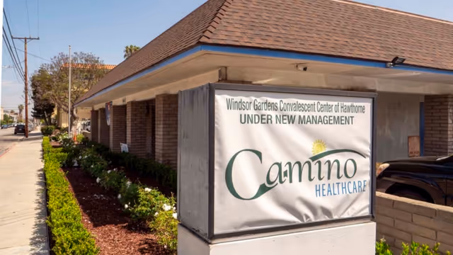 Exterior view of a single-story brick building with a brown shingled roof and a sign in front that reads 'Windsor Gardens Convalescent Center of Hawthorne UNDER NEW MANAGEMENT Camino Healthcare'. There is a sidewalk and some greenery along the side of the building.