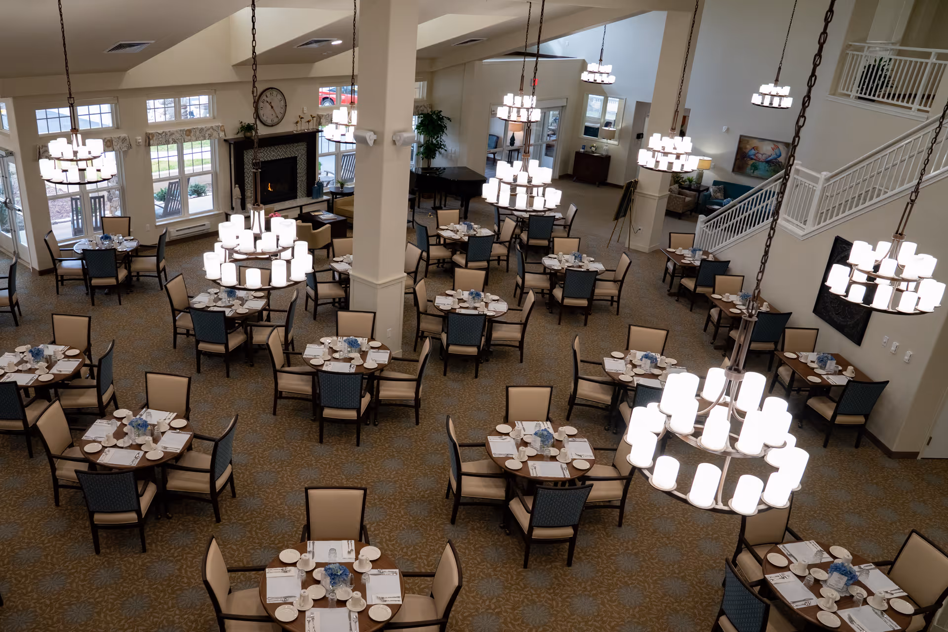 A spacious dining room with multiple round tables set with plates, cups, and napkins. Each table has four chairs with beige and blue upholstery. The room features several large, modern chandeliers hanging from the ceiling, a fireplace with a clock above it, large windows letting in natural light, and a staircase leading to an upper level.