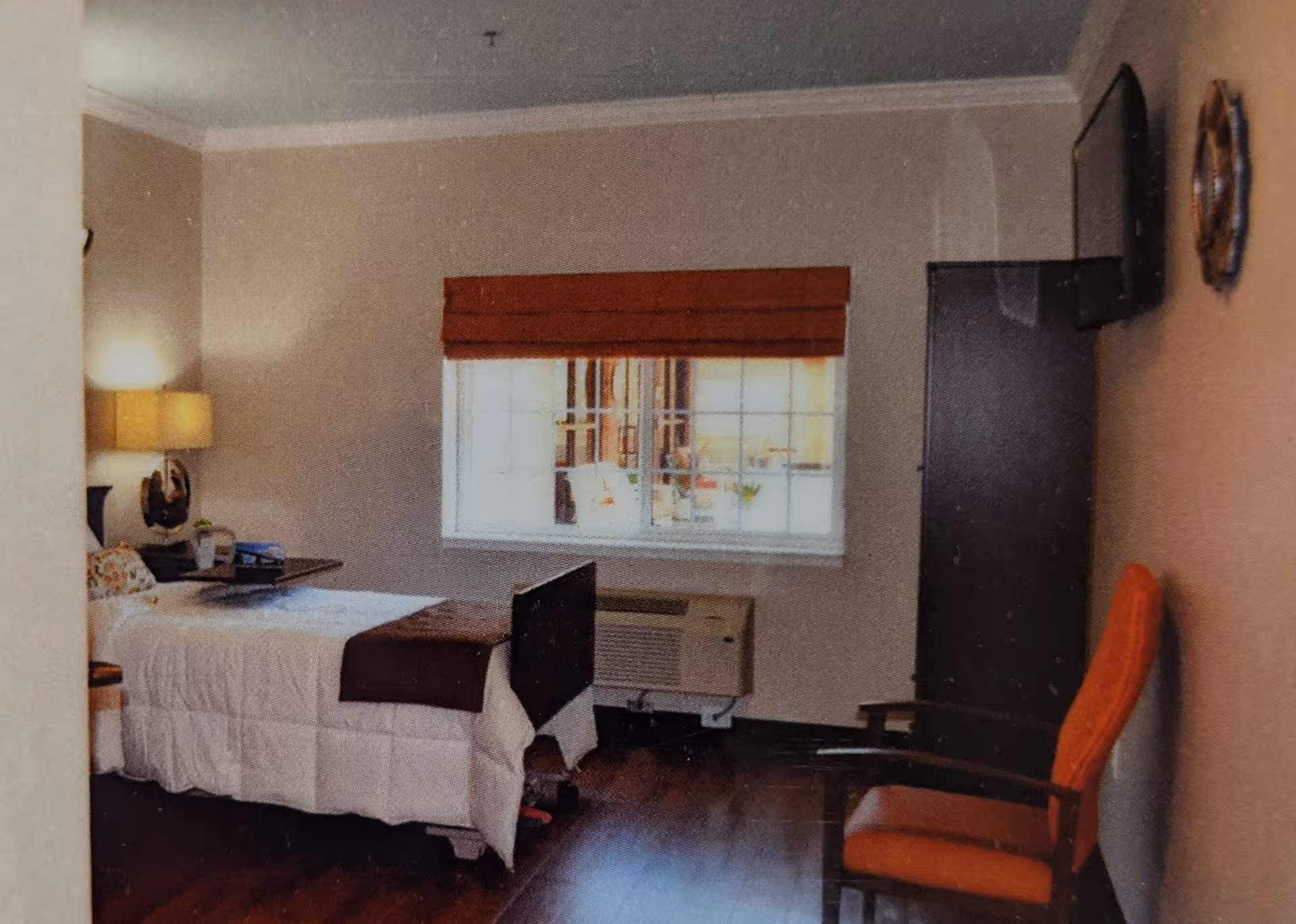 A small bedroom in a healthcare facility featuring a single bed with white bedding and a brown throw, a bedside table with a lamp, a window with a brown blind, an air conditioning unit below the window, a dark wardrobe, a wall-mounted TV, and an orange cushioned chair.